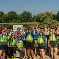 group of 15 in lifejackets standing in front of a lake cheering